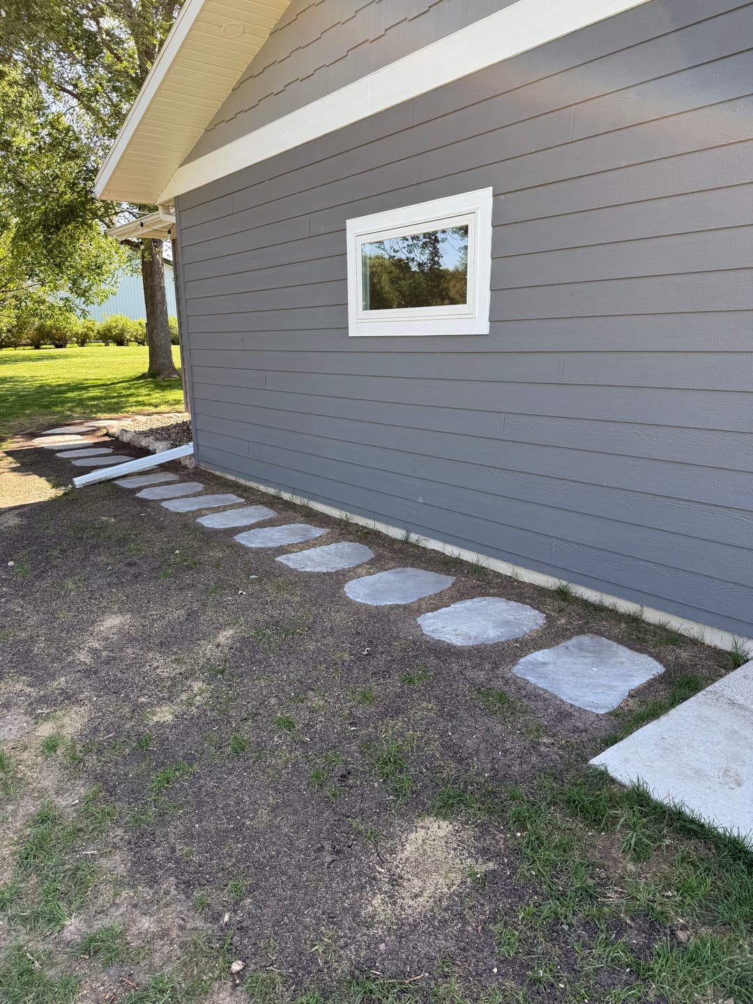 Stepping stones leading to a gray house with a window, surrounded by grass and trees.