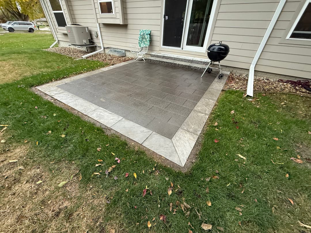Patio with stone pavers and grass, featuring a grill and lawn chair outside a house.