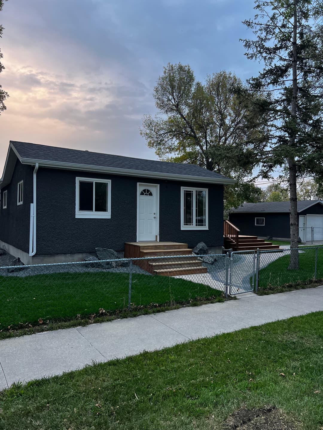 Modern gray house with a white door, surrounded by grass and trees, near a sidewalk.