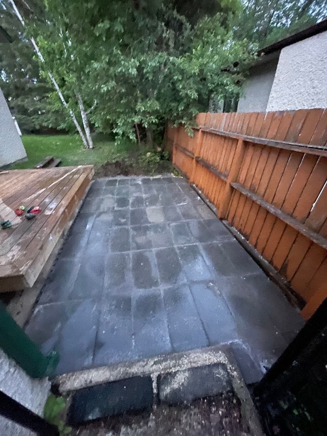 Paved patio area with wooden deck and fence surrounded by greenery.