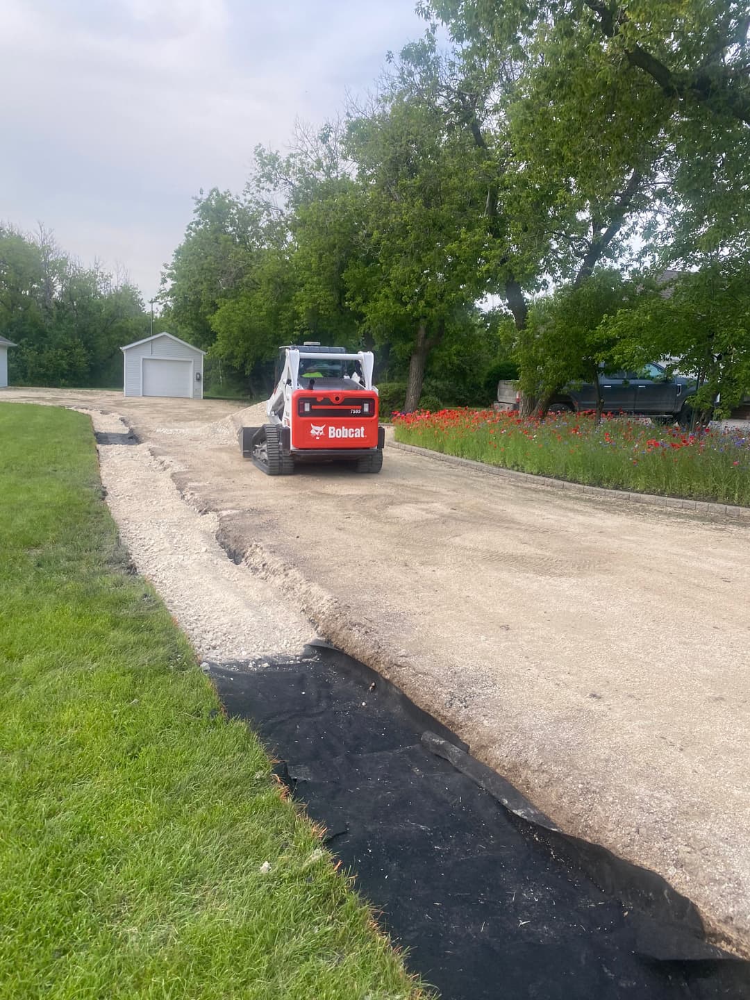 Bobcat skid steer working on gravel road repair, surrounded by lush greenery and flower beds.