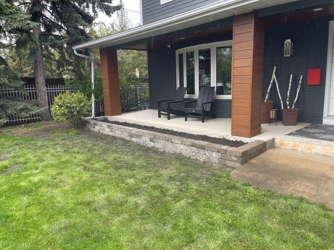 Modern front porch with black chairs, stone wall, and green lawn surrounded by trees.
