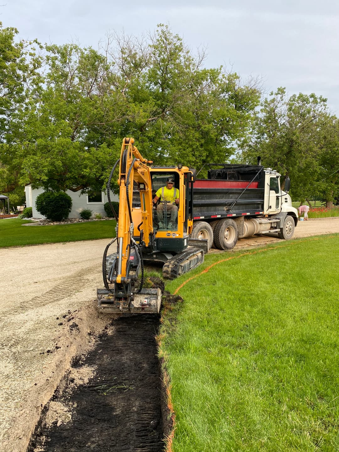 Excavator digging along a gravel road with a dump truck nearby in a residential area.